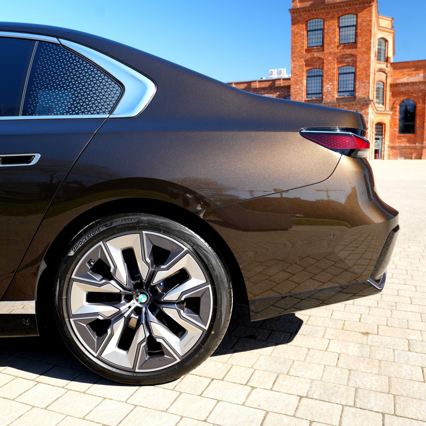 Brązowy samochód BMW z aluminiowymi felgami na tle ceglanego budynku. Widoczny fragment karoserii, tylne światło i wzór na szybie.