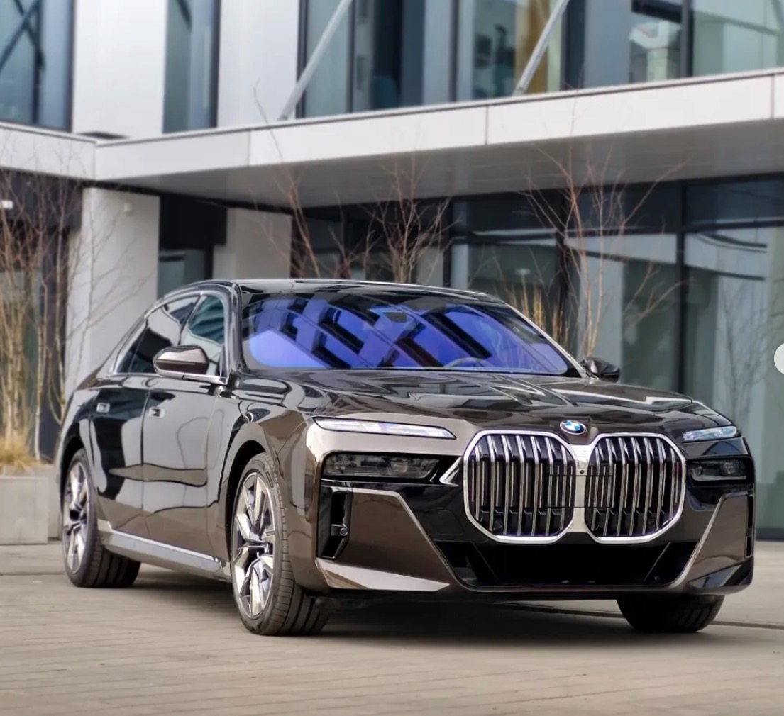 Elegancki, brązowy samochód BMW serii 7 z chromowanym grillem na tle nowoczesnego budynku z przeszklonymi oknami. Luksusowy sedan.