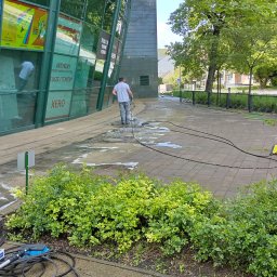 Akcja Renowacja - Mężczyzna czyści kostkę brukową przy budynku biurowym za pomocą myjki ciśnieniowej. Widoczne narzędzia i fragment budynku z reklamami.