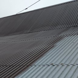 Akcja Renowacja - Częściowo pomalowany, blaszany dach z wyraźnym podziałem na szarą i brązową część. Widoczne złączenia blachy i linia zabezpieczająca na szczycie dachu.