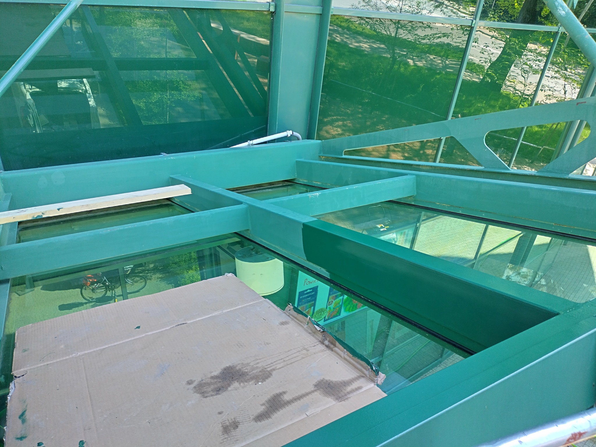 Zielona konstrukcja z szybami, zabezpieczona kartonem. Widoczny rower i zieleń w tle. Prace przygotowawcze do malowania konstrukcji.