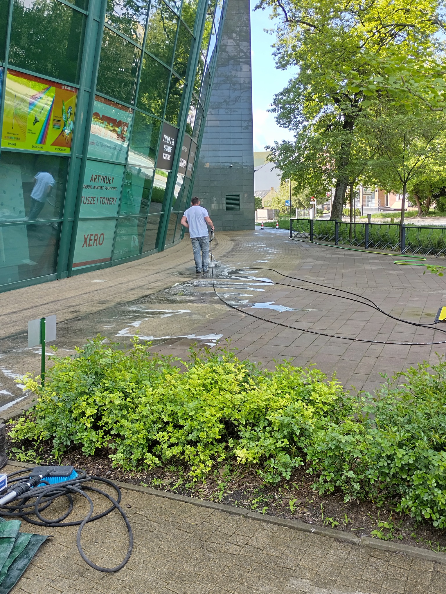 Mężczyzna czyści kostkę brukową przy budynku biurowym za pomocą myjki ciśnieniowej. Widoczne narzędzia i fragment budynku z reklamami.