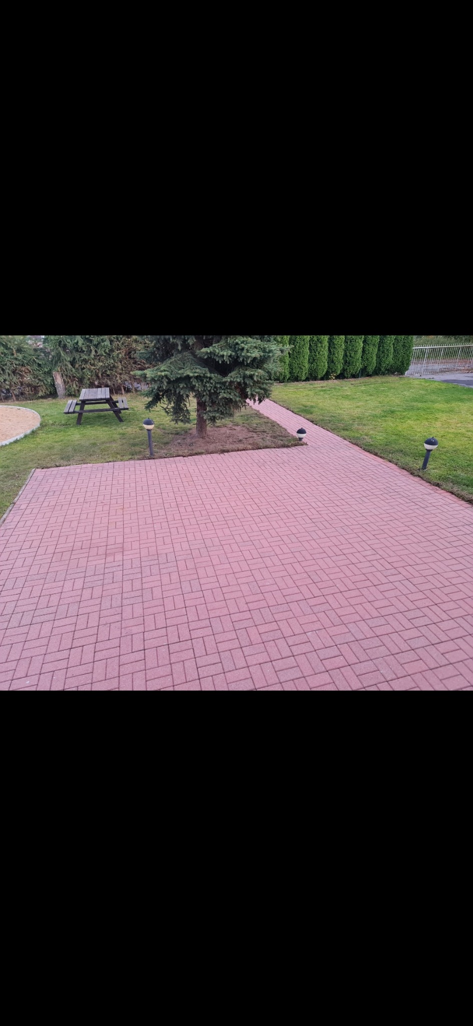 Czerwona kostka brukowa ułożona w jodełkę, ścieżka przez trawnik z lampkami, w tle drzewa iglaste i stół piknikowy. Ujęcie z góry, brak osób.