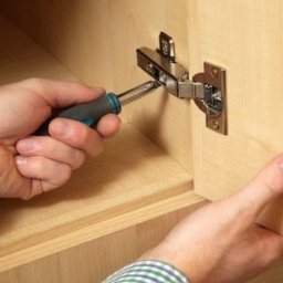 Majster Kazik - Regulacja zawiasu szafki: dłoń trzyma śrubokręt, dopasowując metalowy zawias do drewnianej powierzchni frontu meblowego. Precyzyjne dopasowanie.
