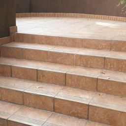 Majster Kazik - Wyłożone beżowymi płytkami schody zewnętrzne prowadzące na taras. Widoczne detale fug i krawędzi płytek. Częściowo widoczna roślinność w tle.