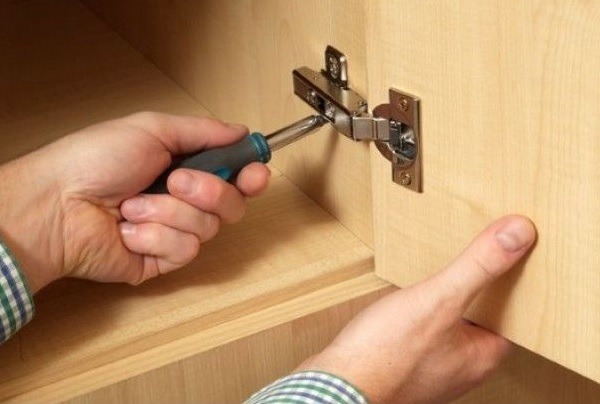 Regulacja zawiasu szafki: dłoń trzyma śrubokręt, dopasowując metalowy zawias do drewnianej powierzchni frontu meblowego. Precyzyjne dopasowanie.