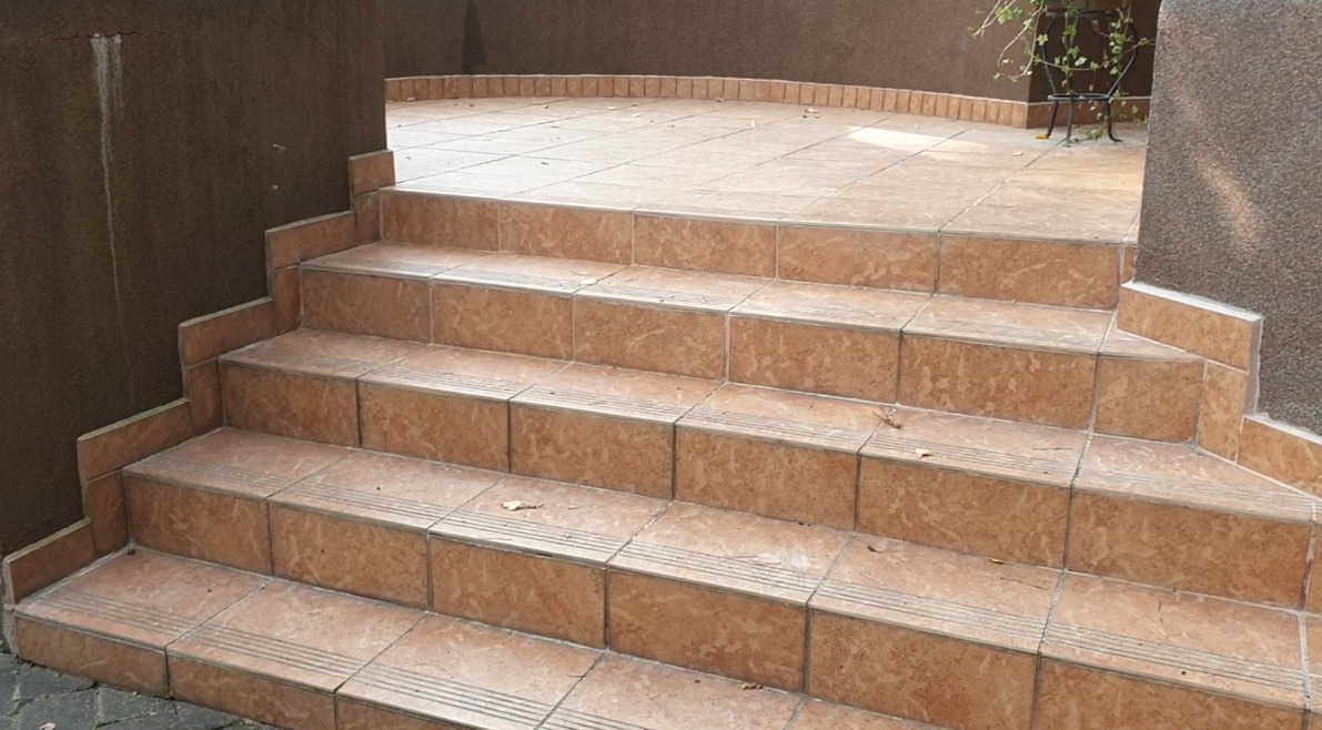 Wyłożone beżowymi płytkami schody zewnętrzne prowadzące na taras. Widoczne detale fug i krawędzi płytek. Częściowo widoczna roślinność w tle.