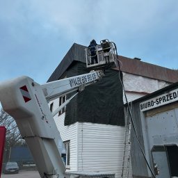Marek Kaczmarski - Prace na wysokości z użyciem podnośnika koszowego, zabezpieczanie budynku czarną plandeką, widoczna drabina oparta o ścianę. Dwie osoby w koszu podnośnika.