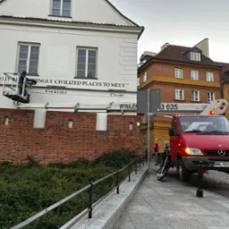 Pracownik w podnośniku koszowym czyści napis 'The Most R...' na fasadzie budynku z ceglaną podstawą. Czerwony samochód z podnośnikiem stoi na chodniku.