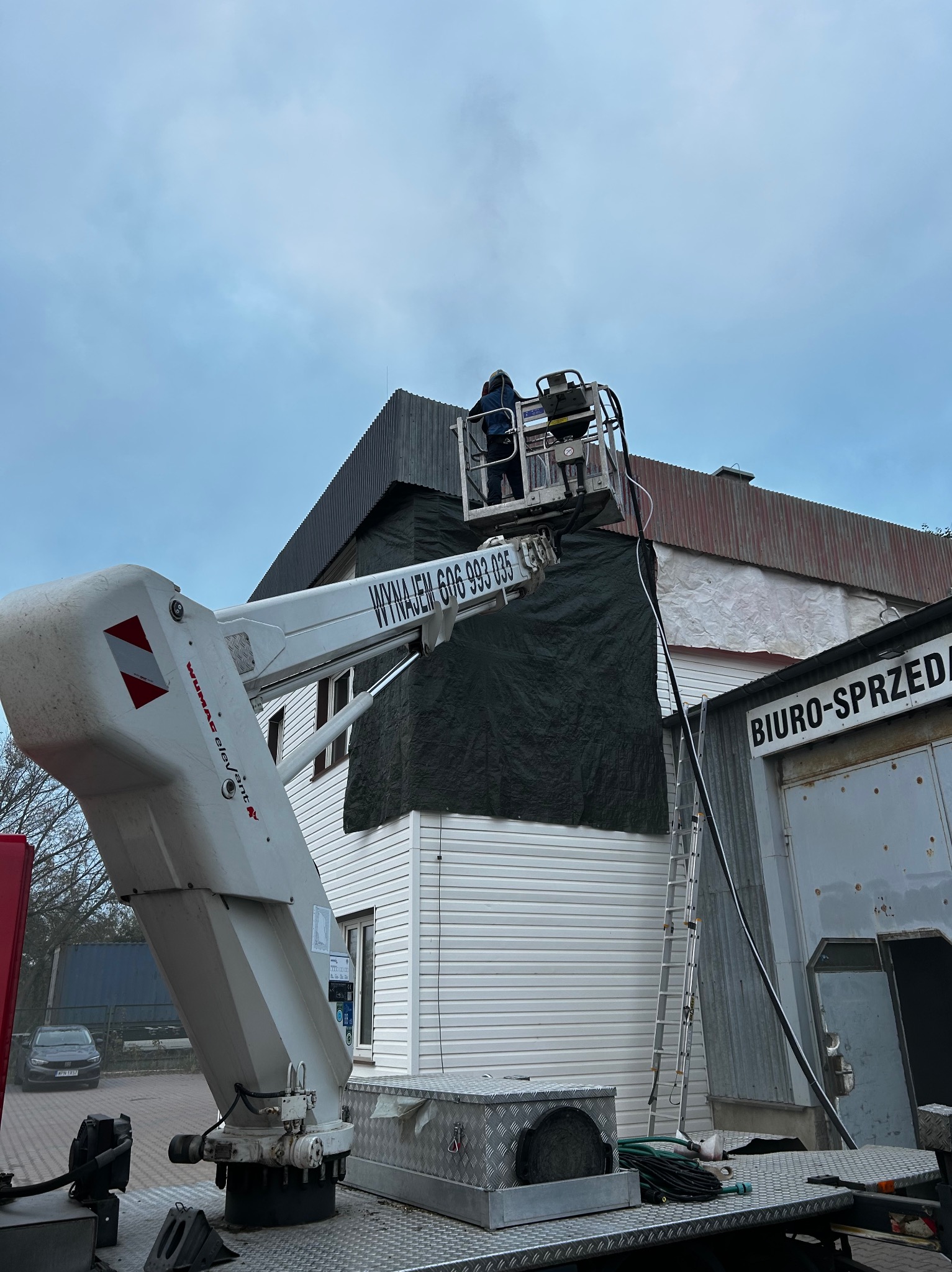 Prace na wysokości z użyciem podnośnika koszowego, zabezpieczanie budynku czarną plandeką, widoczna drabina oparta o ścianę. Dwie osoby w koszu podnośnika.