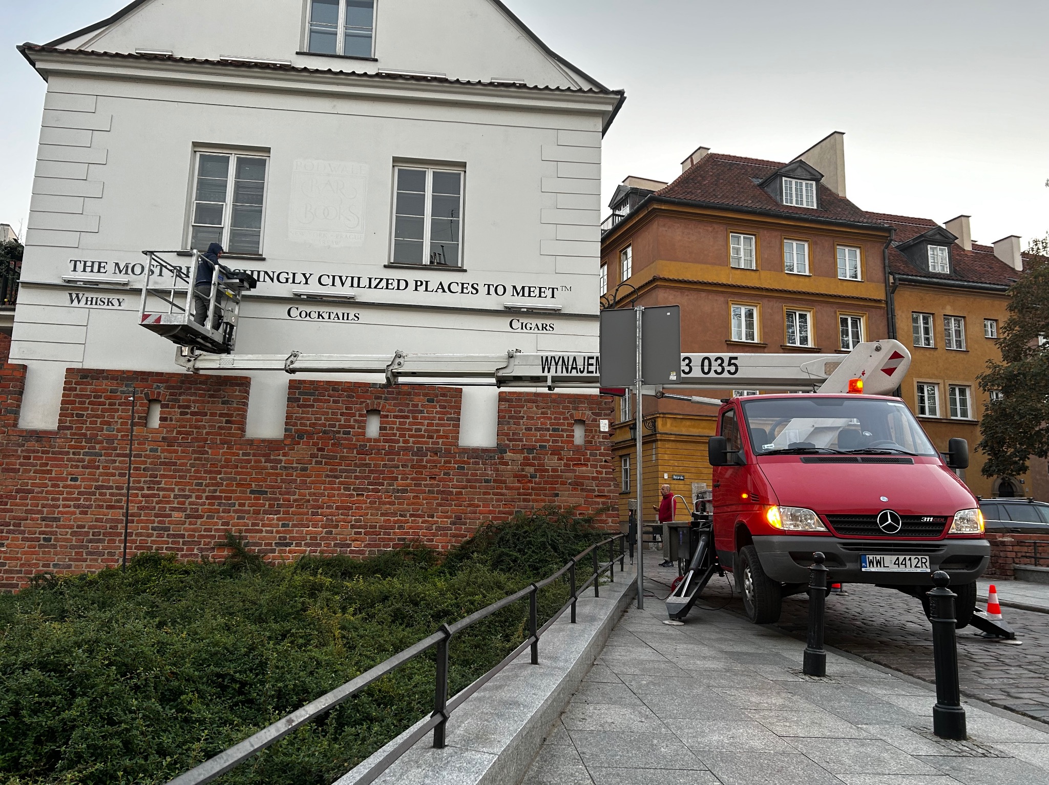Pracownik w podnośniku koszowym czyści napis 'The Most R...' na fasadzie budynku z ceglaną podstawą. Czerwony samochód z podnośnikiem stoi na chodniku.