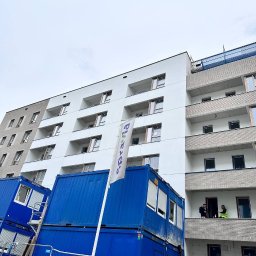 TimBUD - Nowoczesny budynek z białymi balkonami i elewacją, widoczny z dołu, z niebieskimi kontenerami budowlanymi i pracownikami na balkonie.