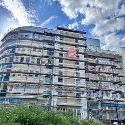 TimBUD - Wielopiętrowy budynek w rusztowaniach pod błękitnym niebem. Na elewacji widoczny baner reklamowy firmy budowlanej. W tle zieleń i fragment chodnika.