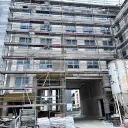 Wielopiętrowy budynek w trakcie prac elewacyjnych, otoczony rusztowaniami. Widoczne materiały budowlane i fragmenty ocieplenia. Ujęcie z dołu, skierowane ku górze.