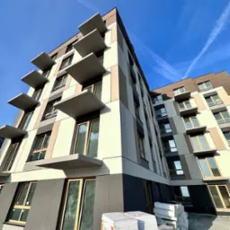 Nowoczesny budynek wielorodzinny z wykończoną elewacją w odcieniach beżu i brązu, z charakterystycznymi, wystającymi balkonami. Na pierwszym planie składowany styropian.