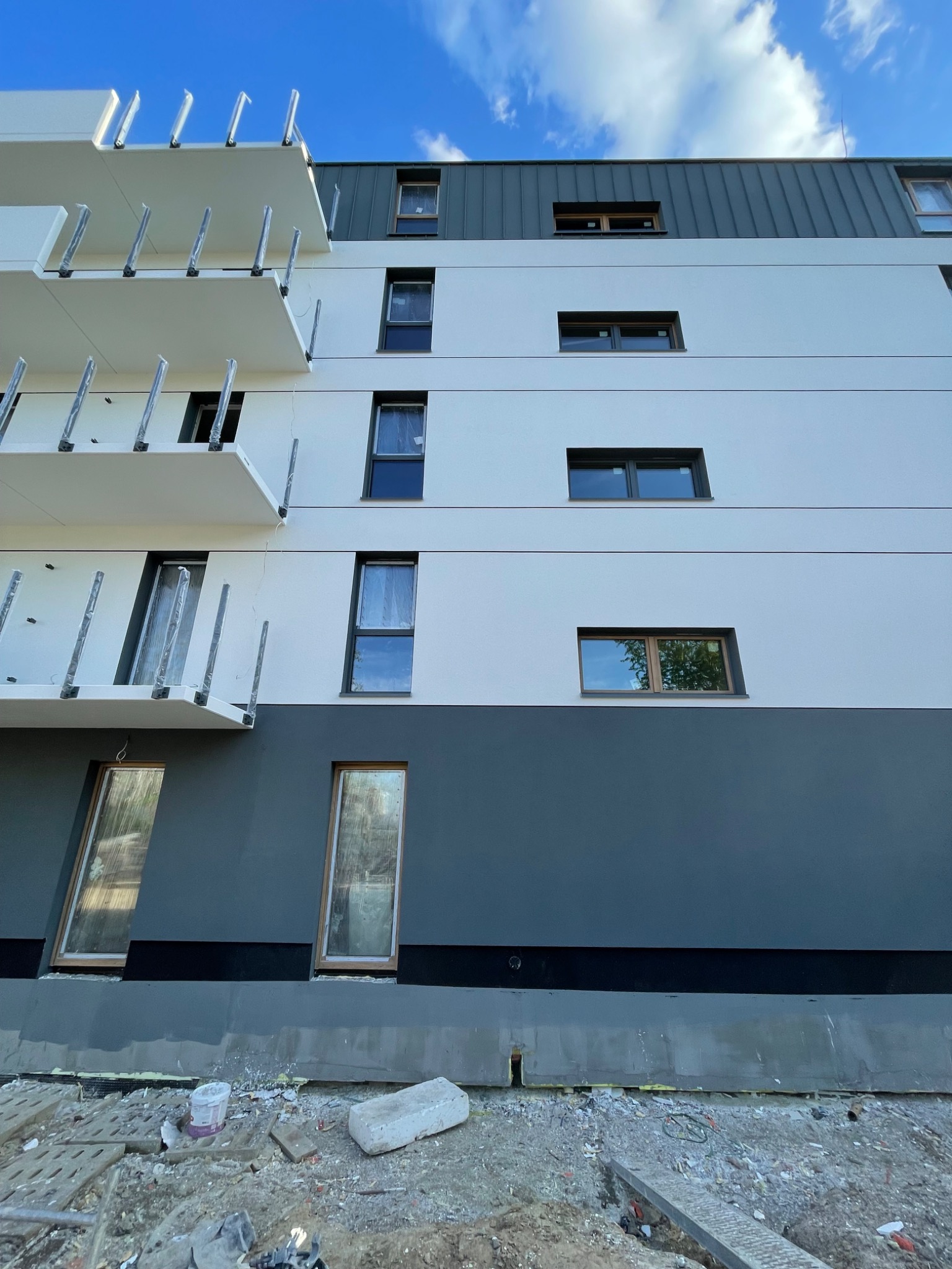 Nowoczesny budynek z ocieploną elewacją w odcieniach bieli i szarości, z widocznymi elementami konstrukcyjnymi balkonów i częściowo zakrytymi oknami folią ochronną.