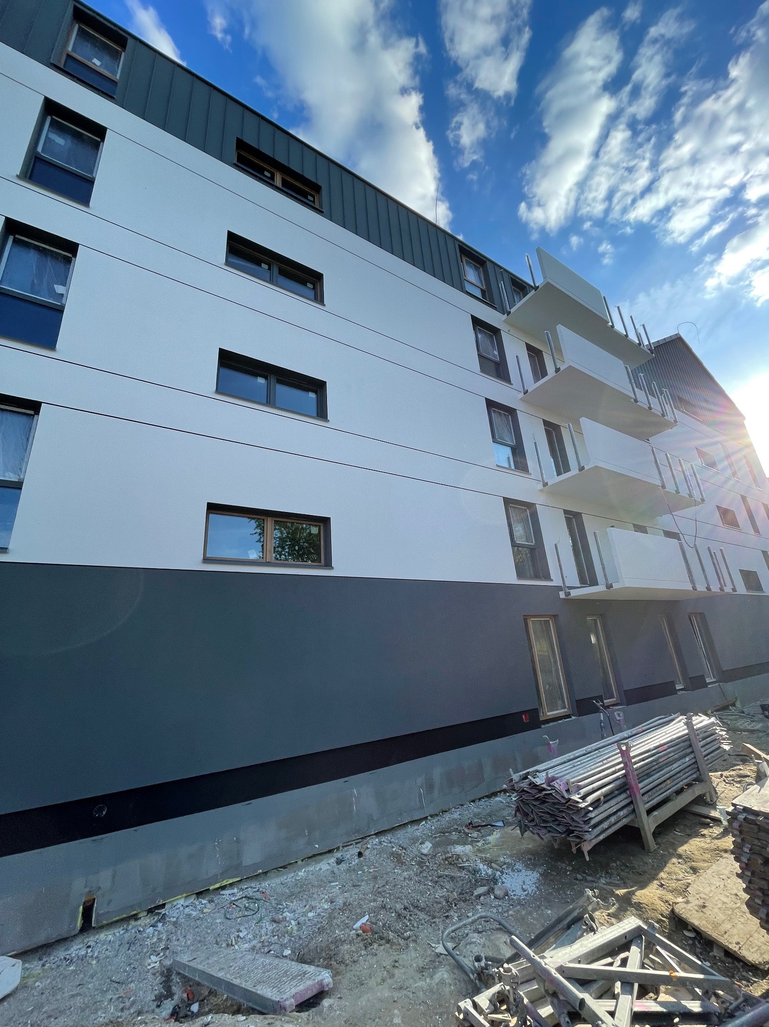 Nowoczesny budynek wielorodzinny z białoszarą elewacją i balkonami, widoczny z dołu pod kątem, z elementami rusztowania na placu budowy w słoneczny dzień.