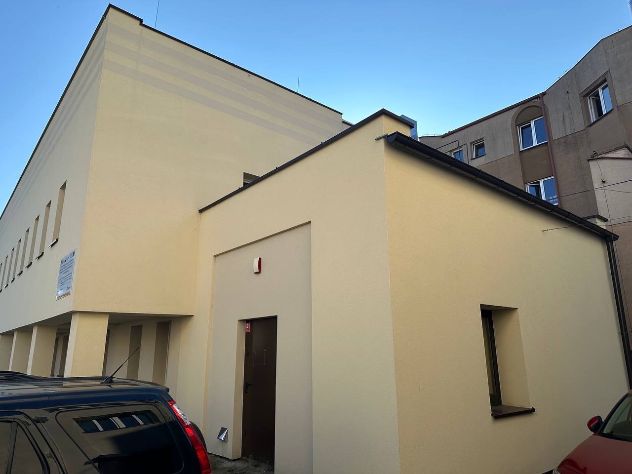 Budynek po renowacji, z jasną elewacją i ciemną stolarką okienną. Widoczne fragmenty zaparkowanych samochodów i sąsiedni budynek. Ujęcie z dołu.