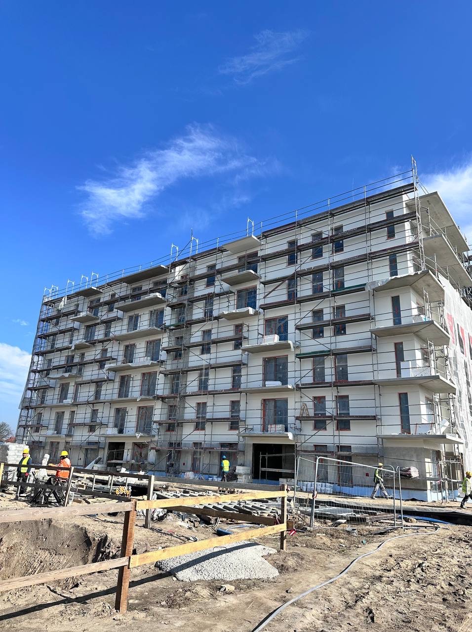 Wielopiętrowy budynek mieszkalny w trakcie prac elewacyjnych, z rusztowaniami i pracownikami w odblaskowych kamizelkach. Błękitne niebo w tle, widok z dołu.