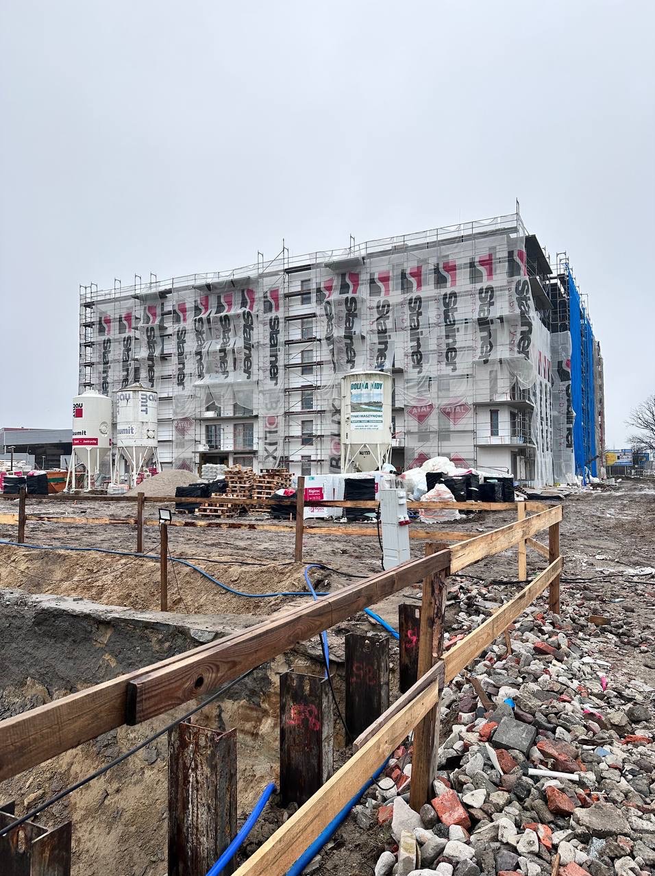 Budynek w rusztowaniach, osłonięty siatką z logotypami, widoczne zbiorniki na materiały budowlane, teren budowy z gruzem i wykopem.