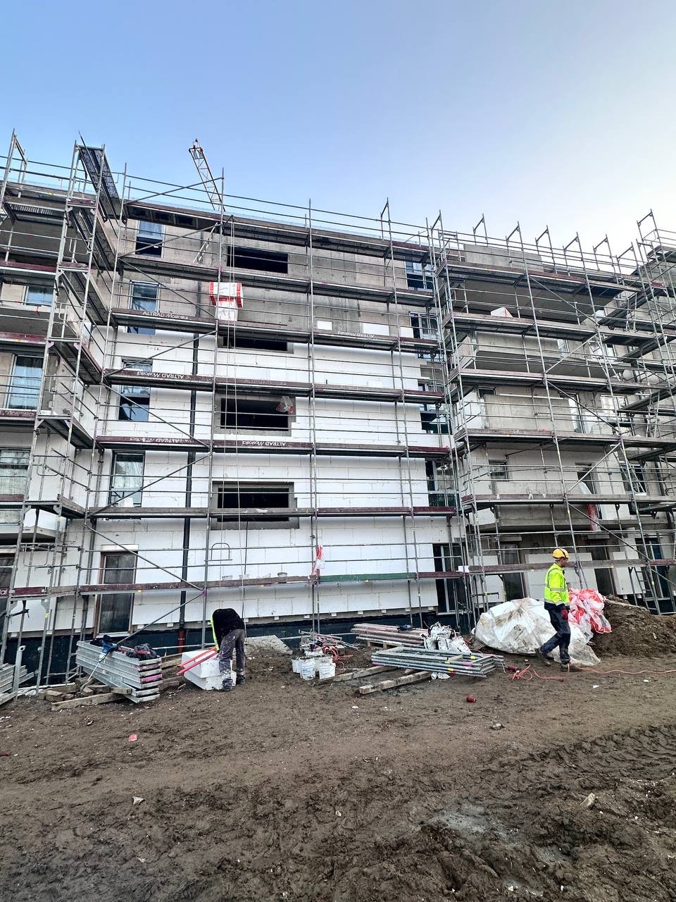 Budynek w trakcie ocieplania, otoczony rusztowaniami. Widoczni pracownicy w odzieży roboczej i materiały budowlane na błotnistym terenie.