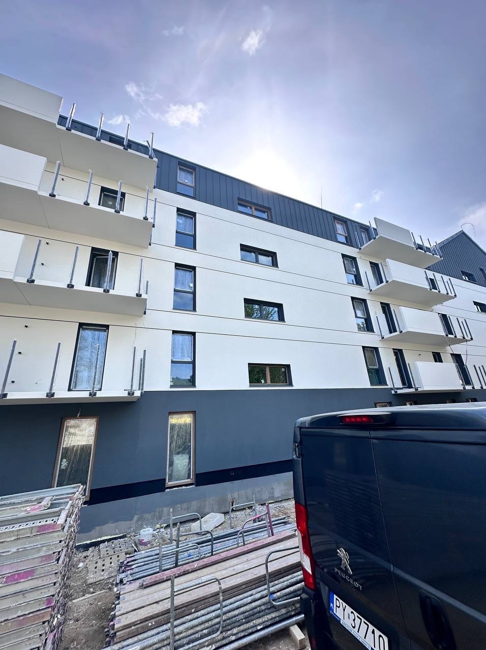 Nowoczesny, pomalowany budynek wielorodzinny z balkonami, z częściowo widocznym samochodem dostawczym i stertą rusztowań. Elewacja w kolorze białym i szarym.