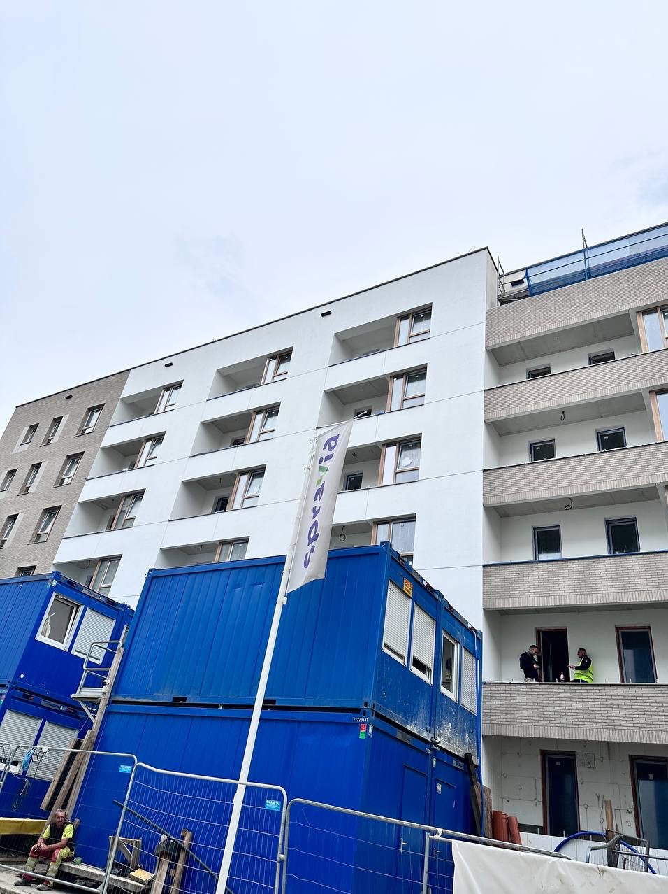 Nowoczesny budynek z białymi balkonami i elewacją, widoczny z dołu, z niebieskimi kontenerami budowlanymi i pracownikami na balkonie.