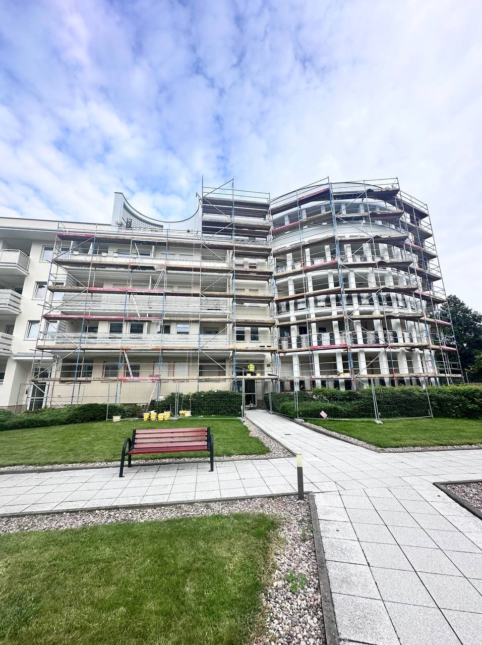 Budynek wielorodzinny w trakcie prac elewacyjnych, otoczony rusztowaniami. Widoczna zieleń i ławka na pierwszym planie, pracownik w żółtym ubraniu na rusztowaniu.