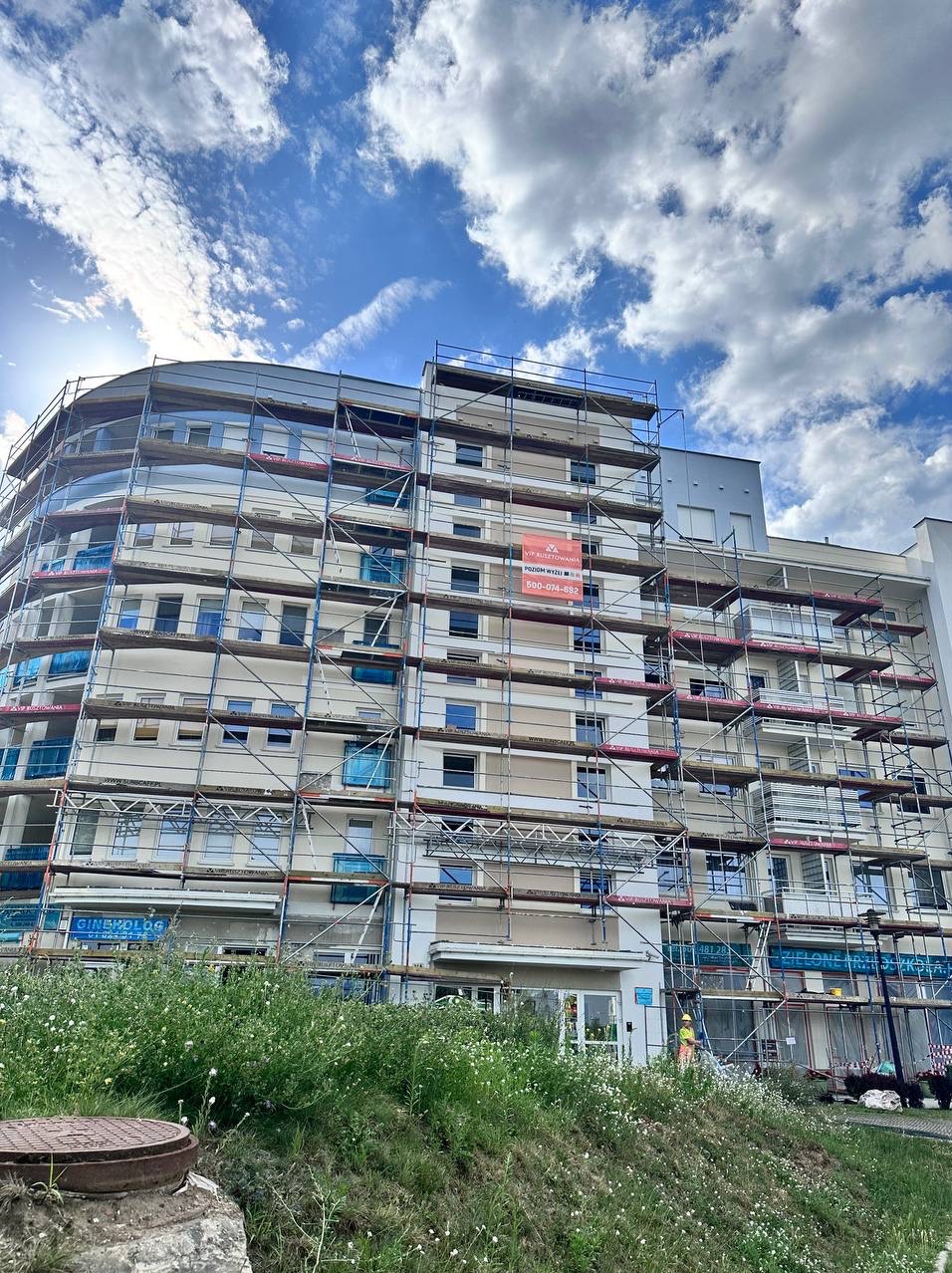Wielopiętrowy budynek w rusztowaniach pod błękitnym niebem. Na elewacji widoczny baner reklamowy firmy budowlanej. W tle zieleń i fragment chodnika.