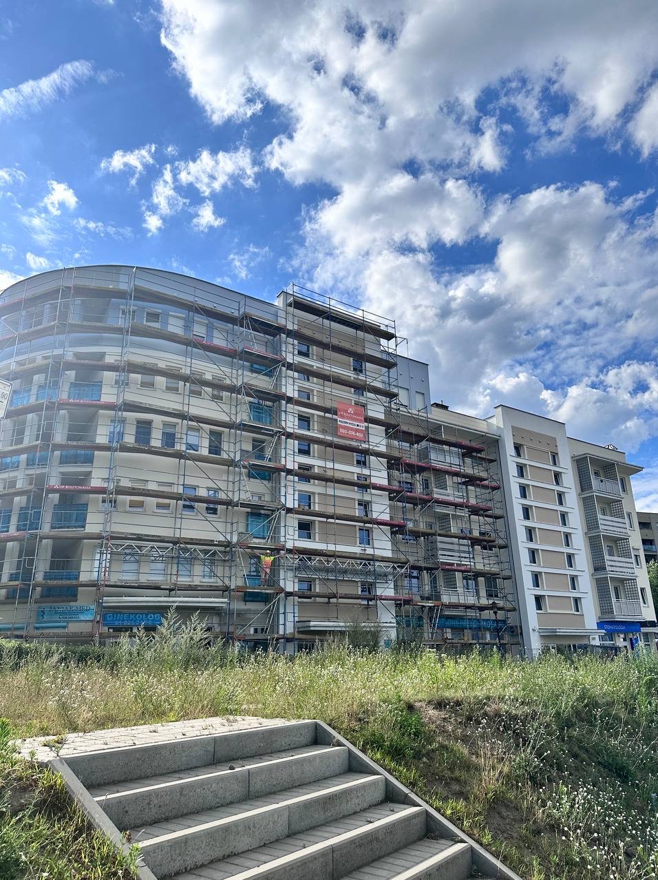 Wielopiętrowy budynek w trakcie prac elewacyjnych, otoczony rusztowaniami. Widoczne schody prowadzące w górę, a w tle błękitne niebo z chmurami.