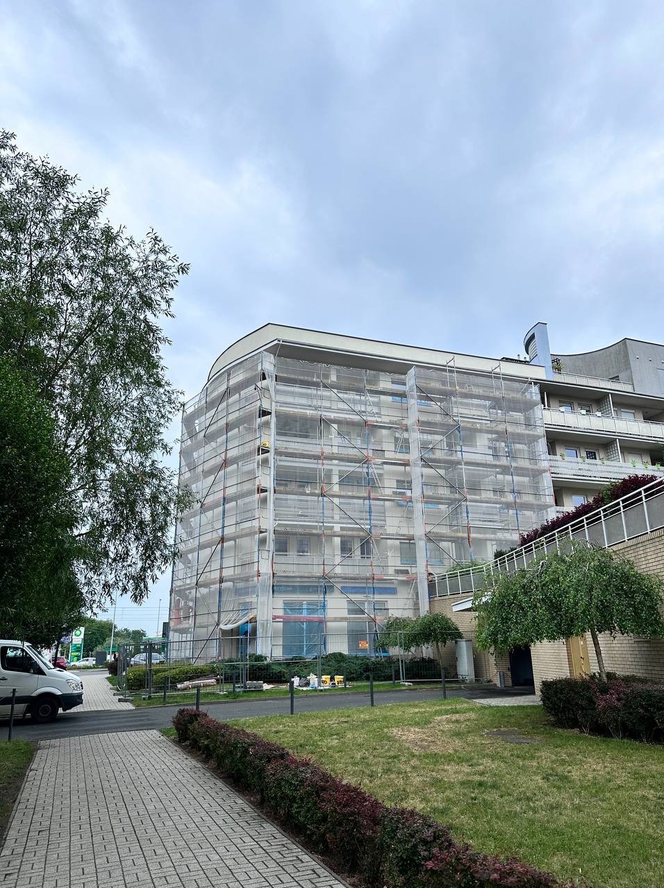 Blok mieszkalny w rusztowaniach, osłonięty siatką zabezpieczającą, widok z poziomu chodnika, zieleń w otoczeniu budynku, pochmurne niebo.