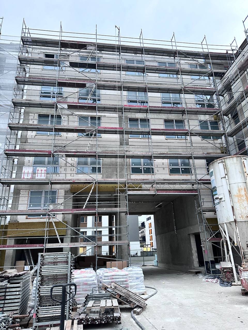 Wielopiętrowy budynek w trakcie prac elewacyjnych, otoczony rusztowaniami. Widoczne materiały budowlane i fragmenty ocieplenia. Ujęcie z dołu, skierowane ku górze.