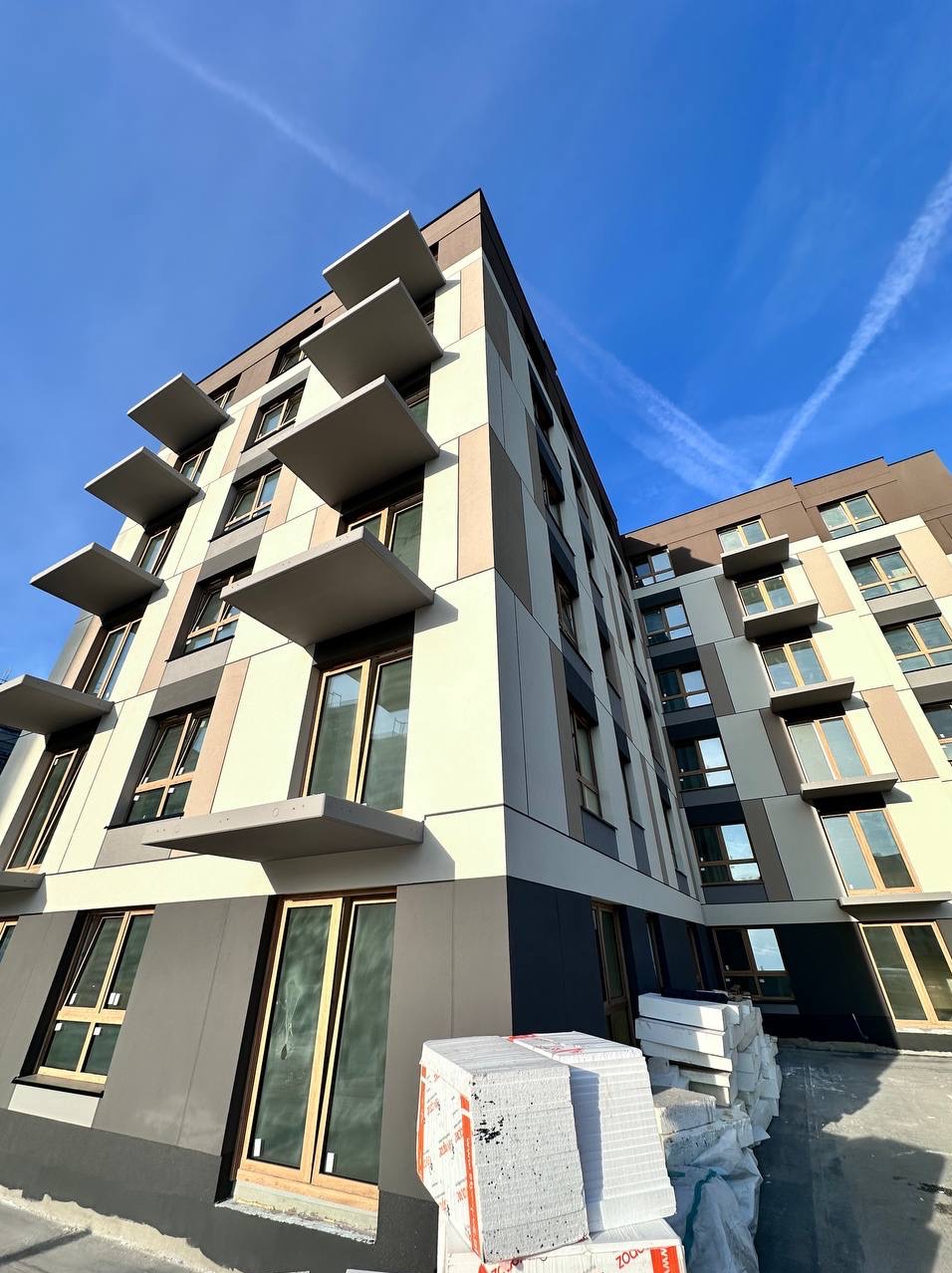 Nowoczesny budynek wielorodzinny z wykończoną elewacją w odcieniach beżu i brązu, z charakterystycznymi, wystającymi balkonami. Na pierwszym planie składowany styropian.