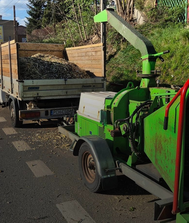 Ciężarówka z przyczepionym rębakiem do gałęzi, wypełniona zrębkami. Widoczne tablice rejestracyjne pojazdów. Praca w toku, na asfalcie leżą zrębki.