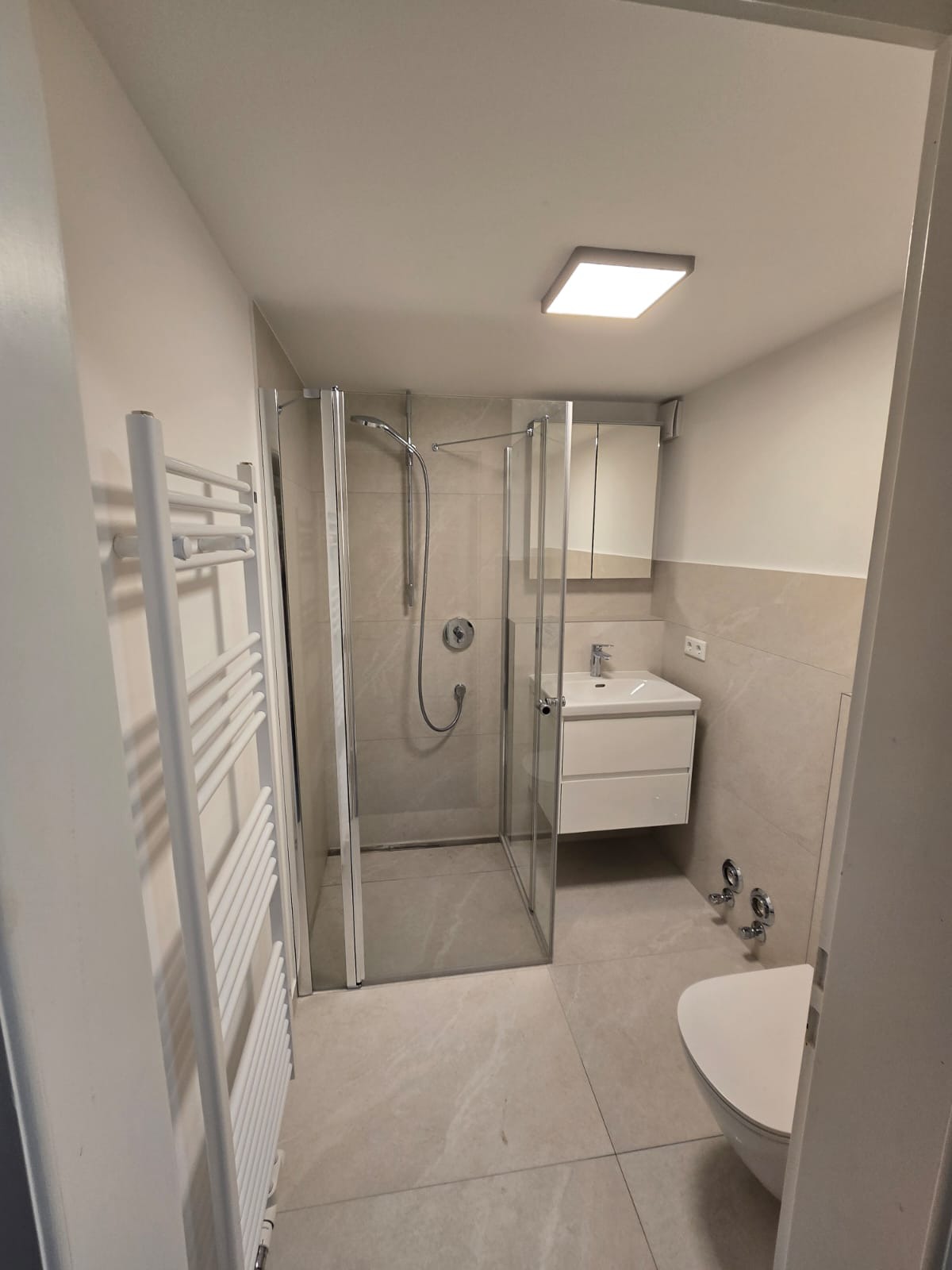 Jasna łazienka po remoncie: kabina prysznicowa ze szklanymi drzwiami, biała umywalka z szafką, grzejnik drabinkowy. Neutralna kolorystyka płytek i armatury.