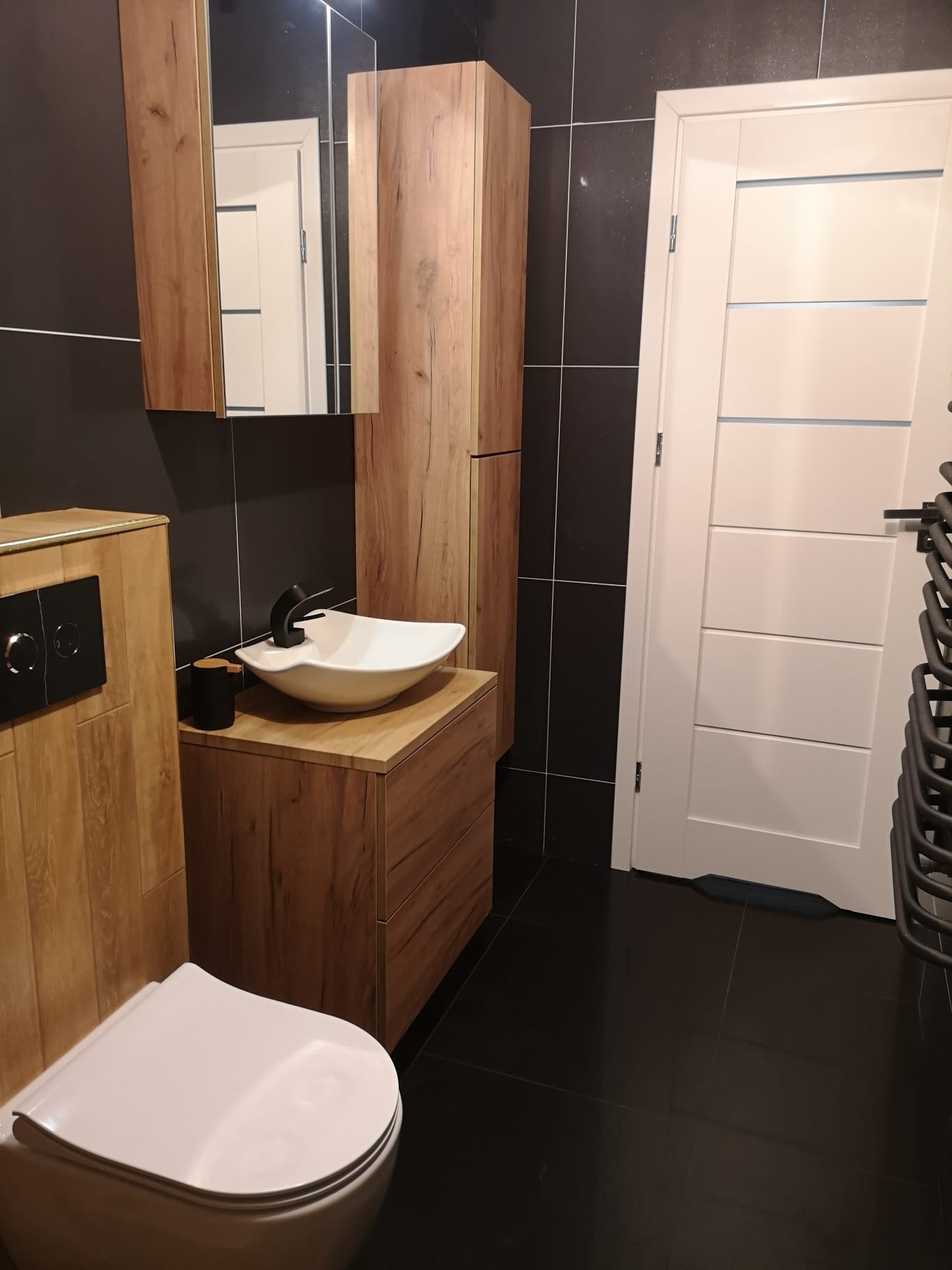 Nowoczesna łazienka z drewnianymi szafkami, białą umywalką i czarnymi płytkami. Minimalistyczny design, ciepłe oświetlenie, eleganckie wykończenie.