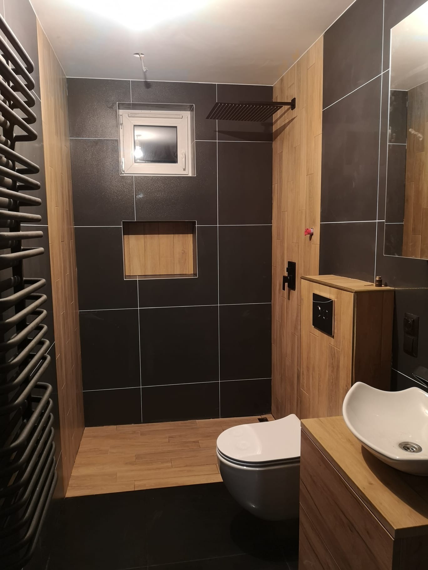 Nowoczesna łazienka z czarnymi płytkami i drewnopodobnymi akcentami. Umywalka nablatowa, WC podwieszane, grzejnik drabinkowy i deszczownica.