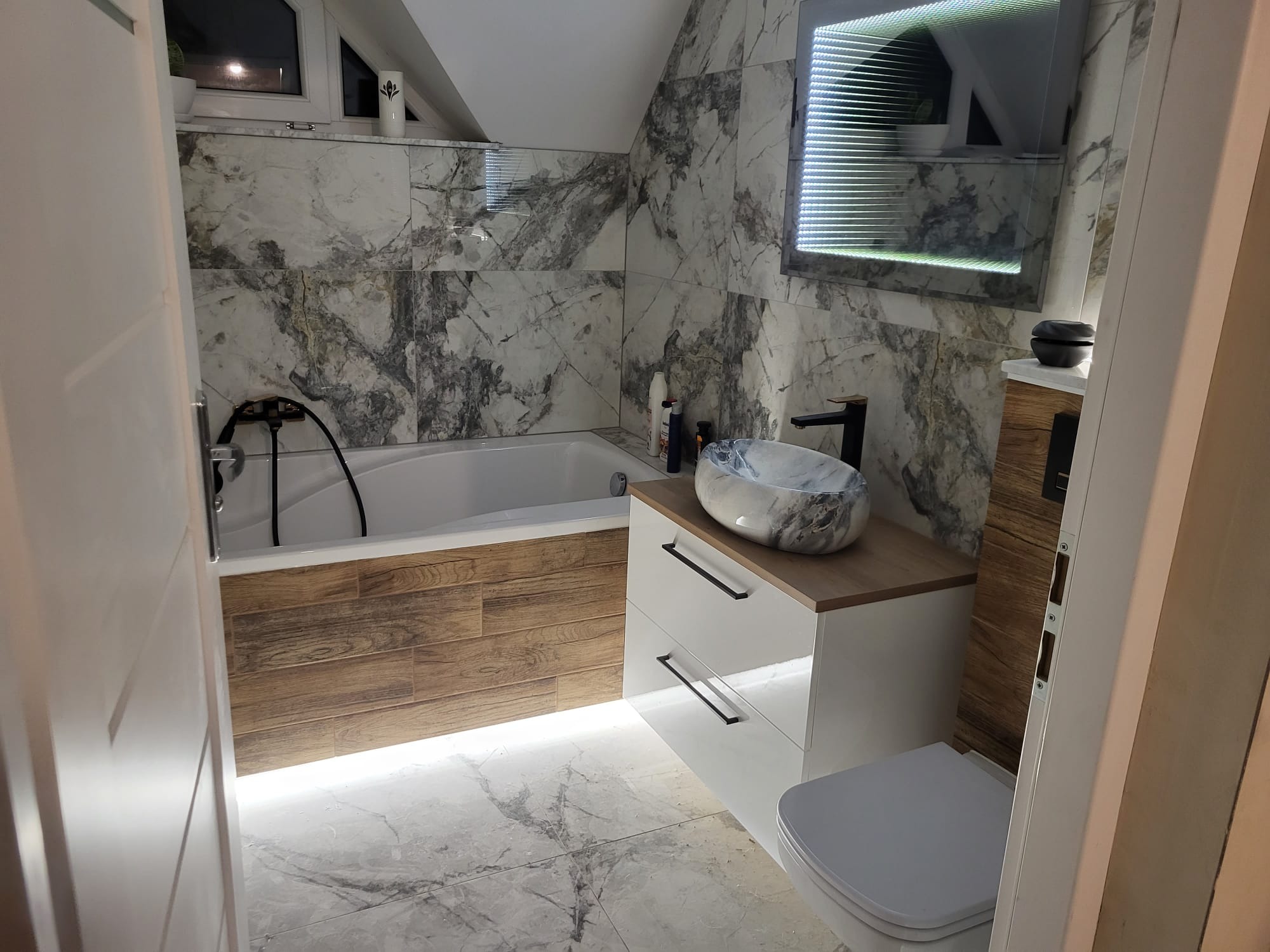 Elegancka łazienka na poddaszu z marmurowymi płytkami, podświetleniem LED pod wanną i szafką, oraz oryginalną umywalką. Nowoczesne wykończenie w jasnych barwach.