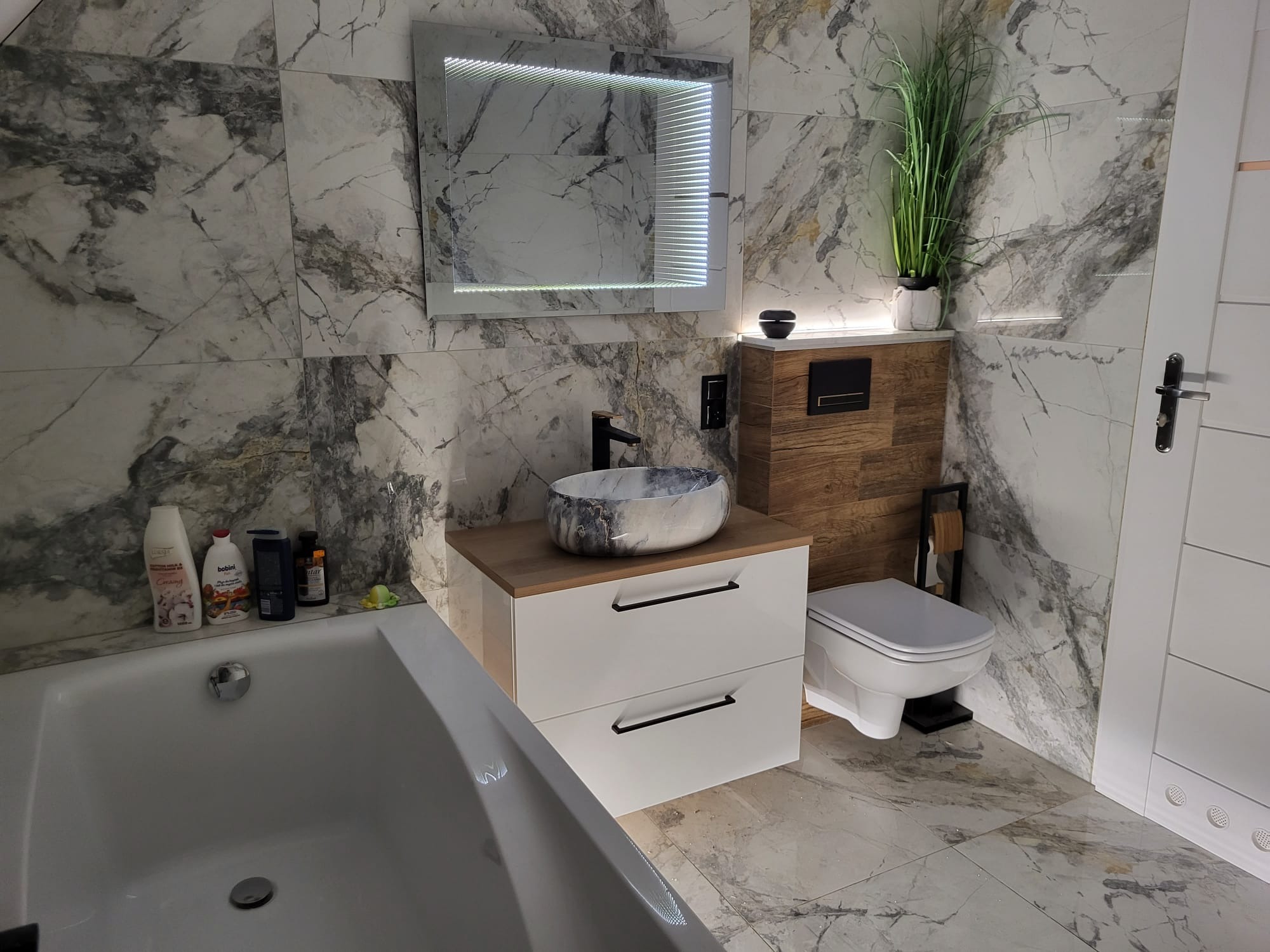 Wykończona łazienka z marmurowymi płytkami, podświetlanym lustrem i umywalką nablatową. Widoczna wanna, toaleta i szafka z szufladami.
