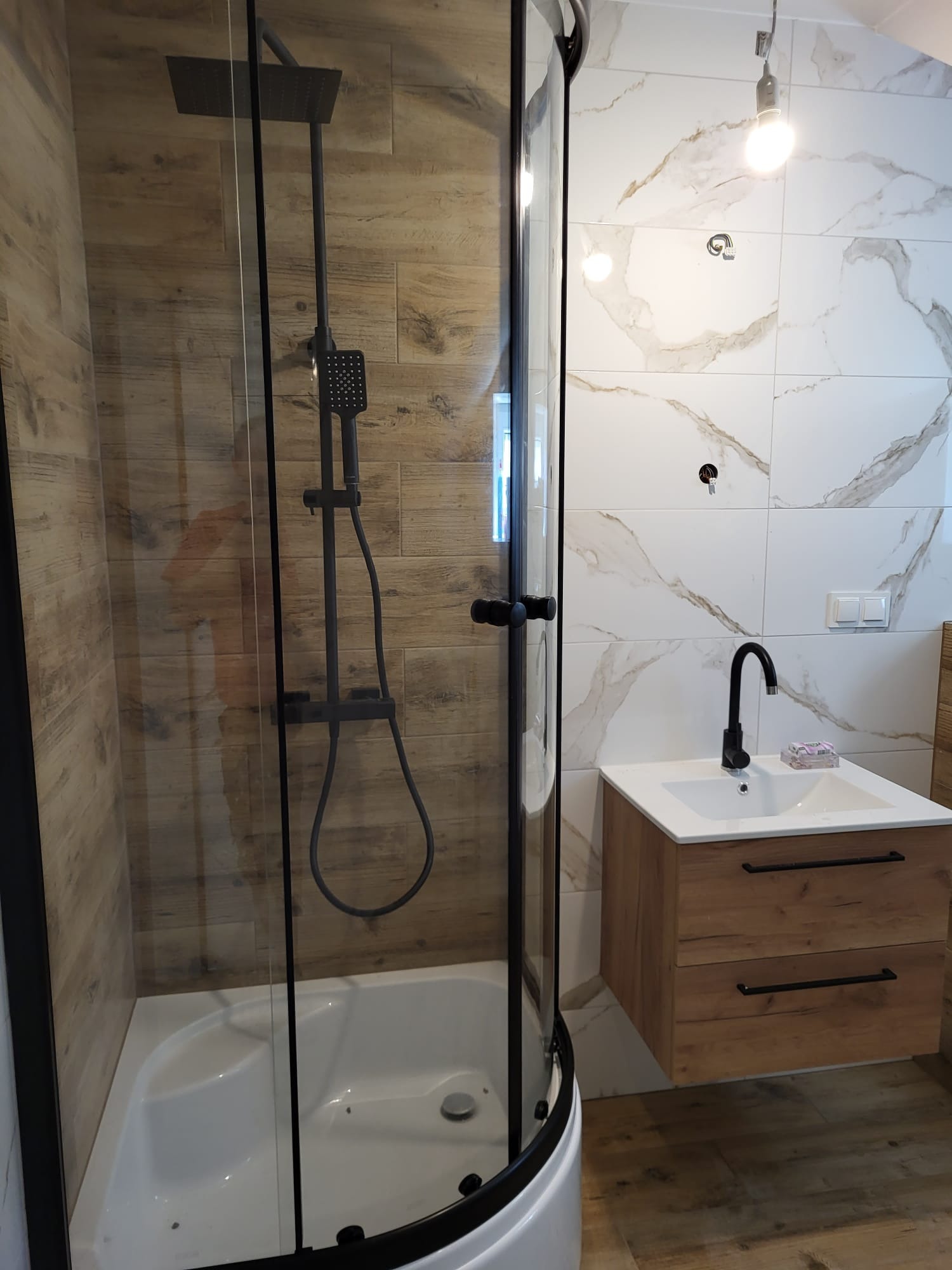Nowoczesna łazienka z kabiną prysznicową typu walk-in, umywalką z szafką w odcieniach drewna oraz białymi płytkami imitującymi marmur. Czarna armatura.