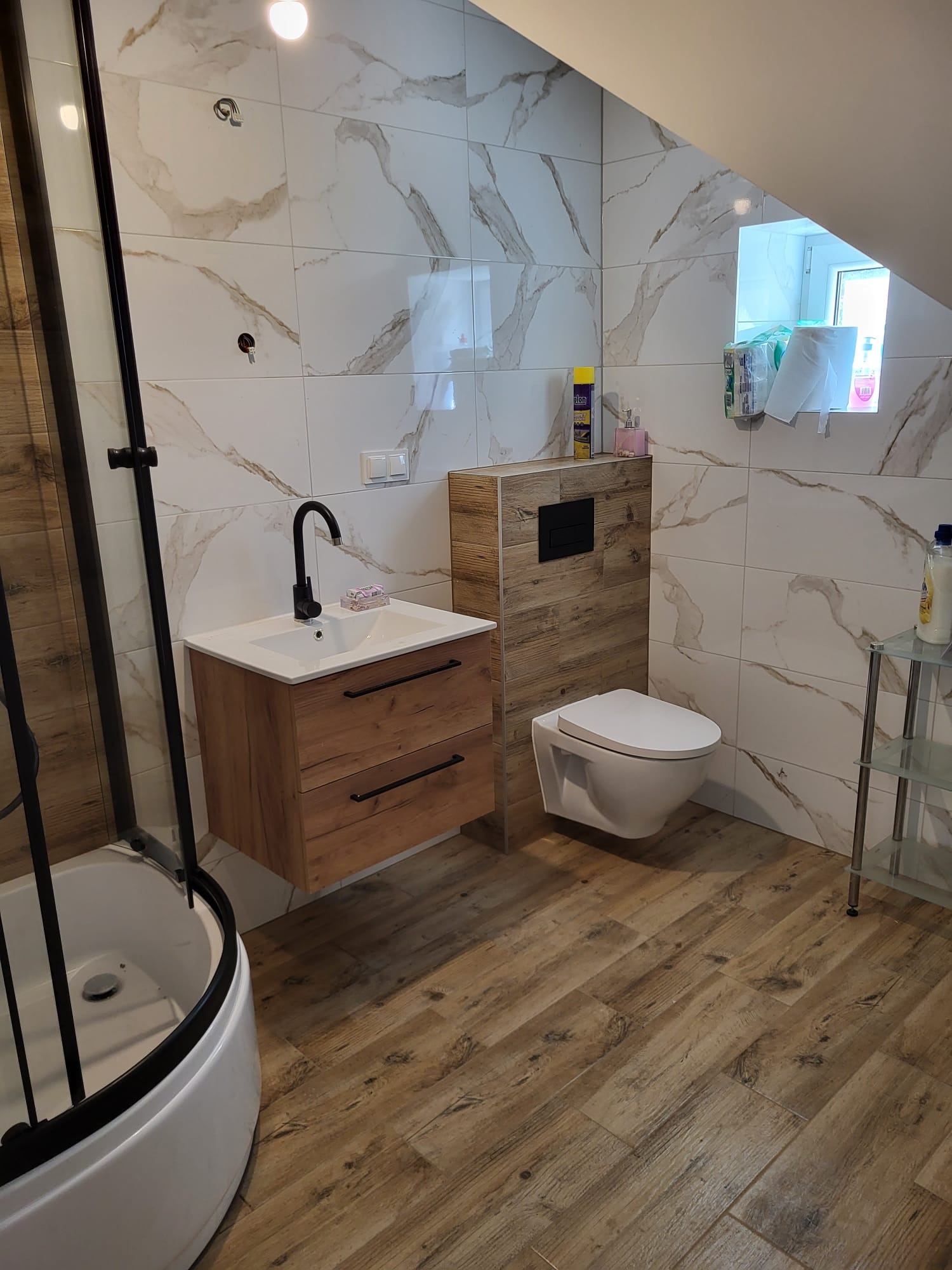 Ukończona łazienka z kabiną prysznicową, umywalką z drewnianą szafką, podwieszaną toaletą i marmurowymi płytkami na ścianach i podłodze, oświetlona światłem dziennym.