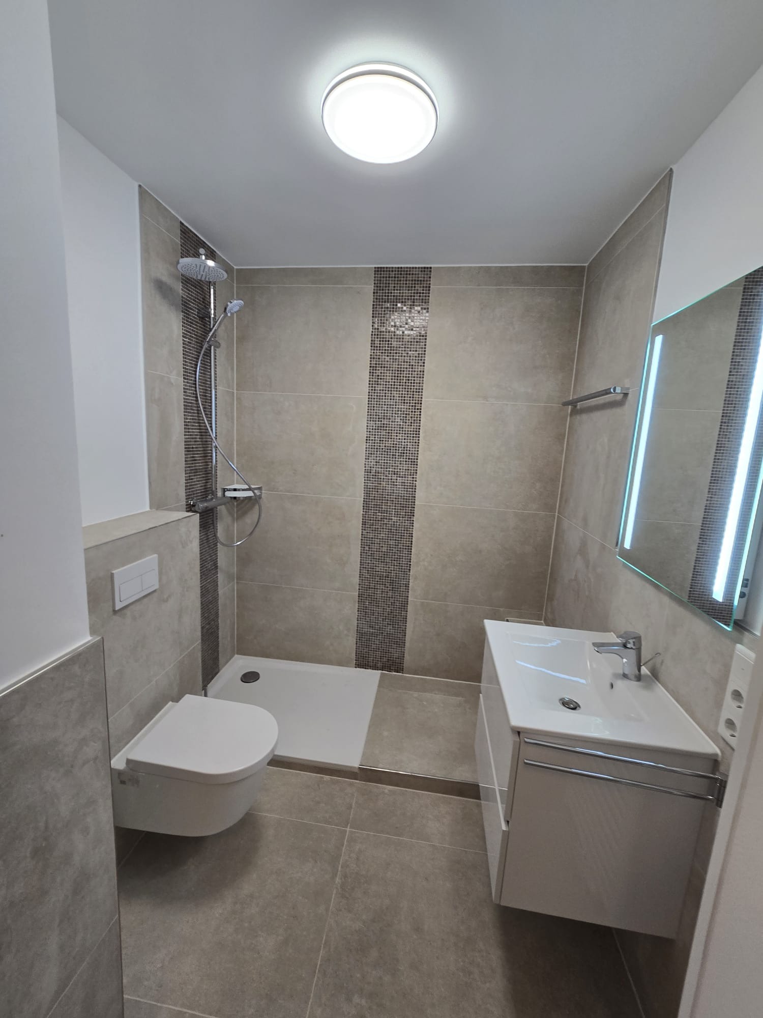 Wykończona łazienka z kabiną prysznicową, umywalką z szafką i podwieszaną toaletą. Płytki w odcieniach beżu i szarości z dekoracyjnym pasem mozaiki. Oświetlenie sufitowe.