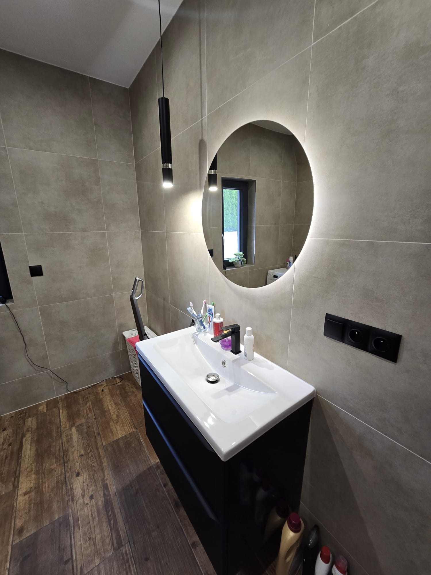 Nowoczesna łazienka z drewnopodobną podłogą, szarą glazurą i okrągłym lustrem z podświetleniem LED. Widoczna czarna szafka pod umywalką i minimalistyczna lampa.