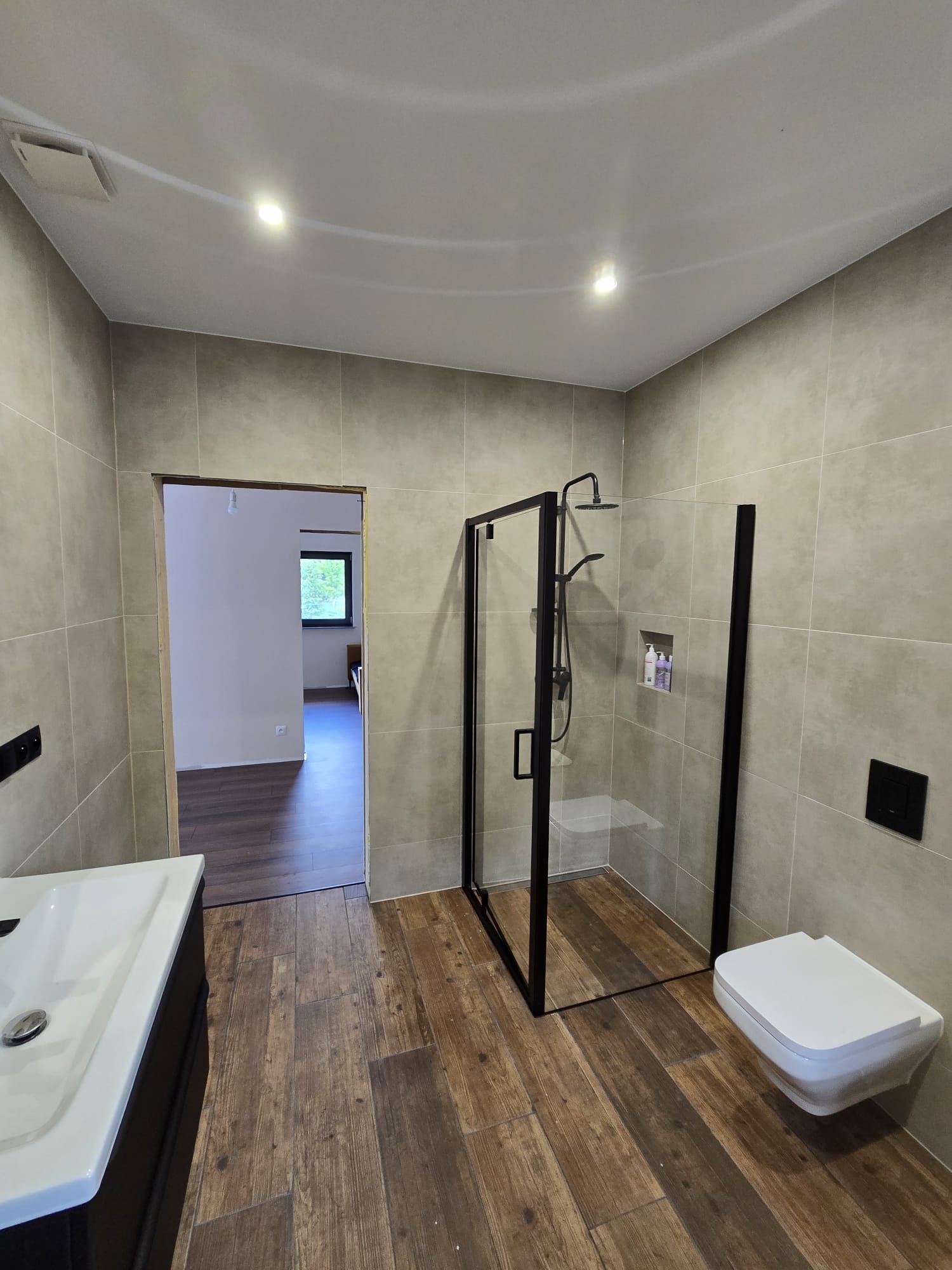 Wykończona łazienka z kabiną prysznicową typu walk-in z czarnymi ramami, wiszącą toaletą i umywalką. Na podłodze drewnopodobne płytki, na ścianach jasnoszare płytki.