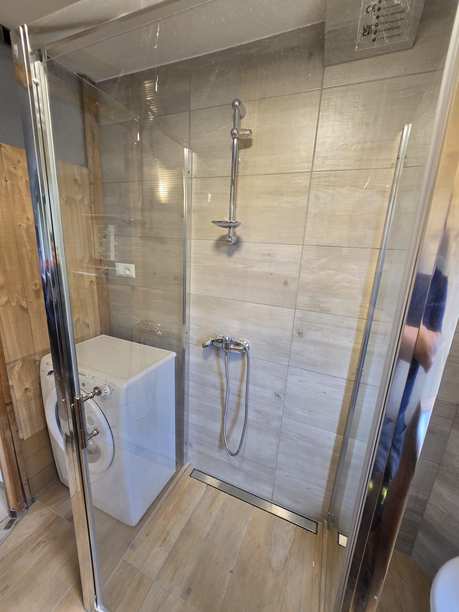 Kabina prysznicowa z deszczownicą i odpływem liniowym, jasne płytki imitujące drewno, pralka w tle, nietypowe rozwiązanie aranżacyjne małej łazienki.