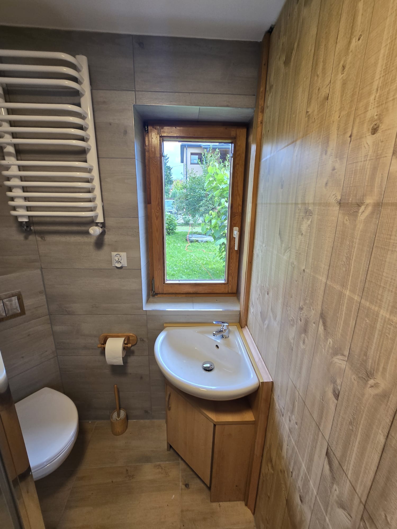 Jasna łazienka z oknem: drewnopodobne płytki na ścianie, biała umywalka z szafką, grzejnik drabinkowy i widok na ogród przez okno.