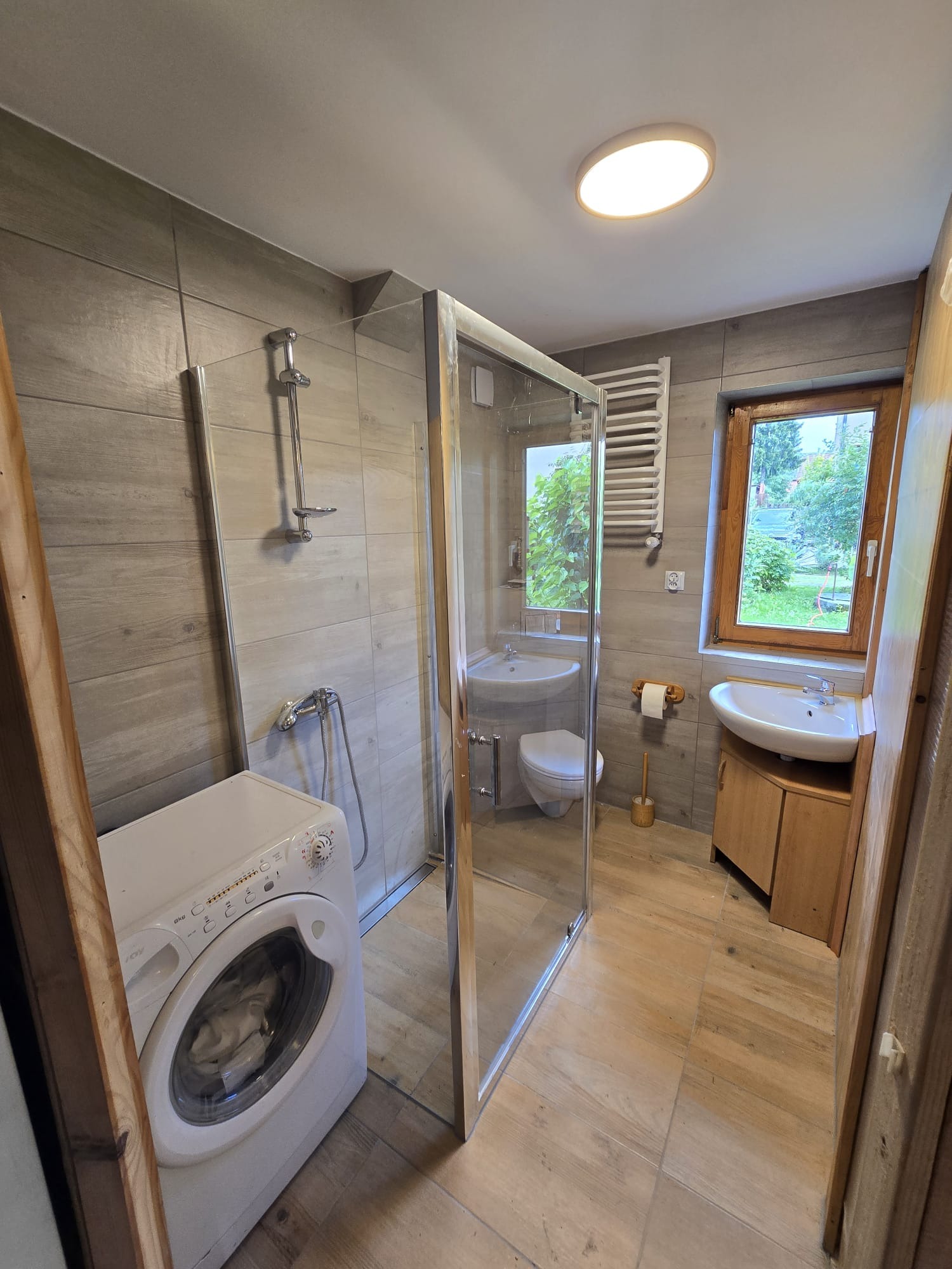 Jasna łazienka z kabiną prysznicową, pralką i dwoma umywalkami. Płytki imitujące drewno na podłodze i ścianach. Okno z widokiem na zieleń.