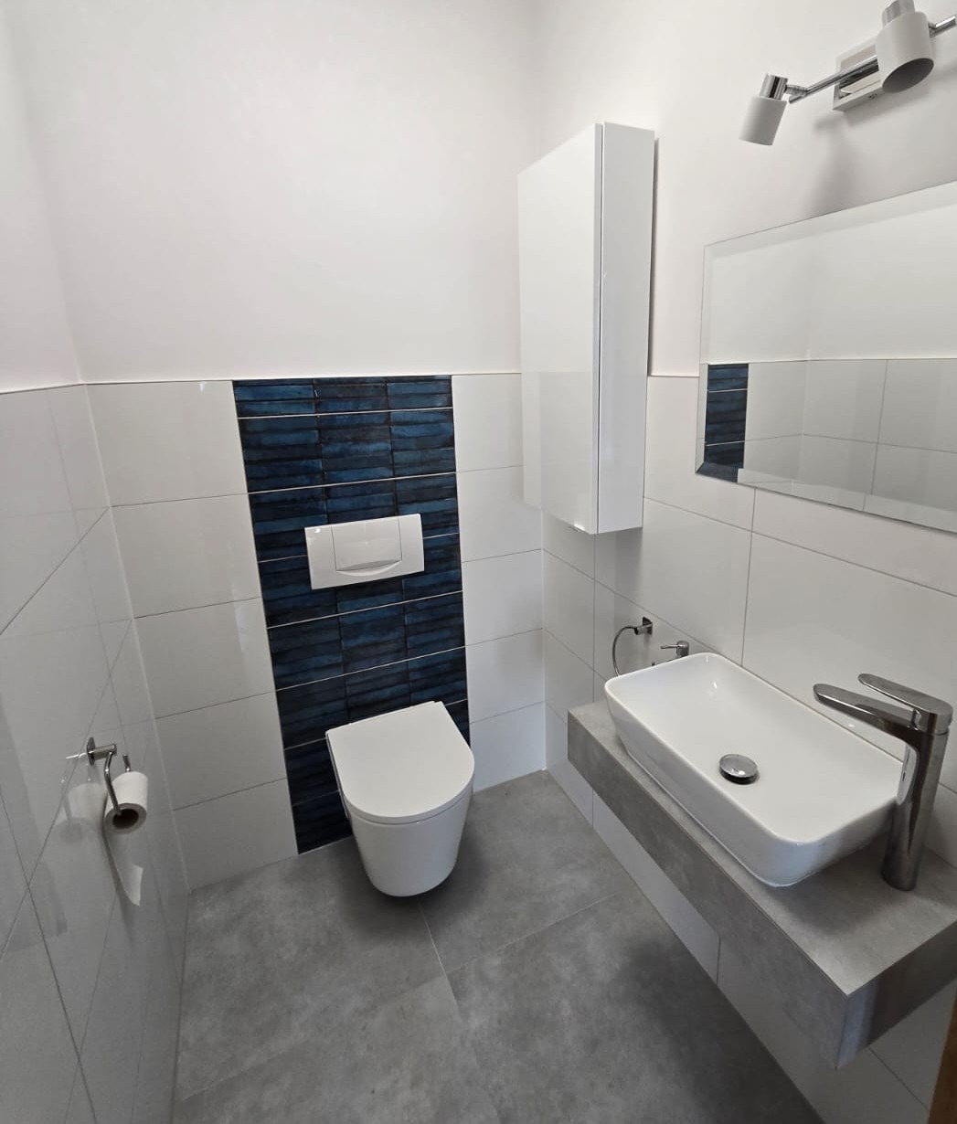 Nowoczesna łazienka po remoncie: biało-szare płytki, umywalka nablatowa na betonowym blacie, podwieszana toaleta i akcent z granatowych płytek.