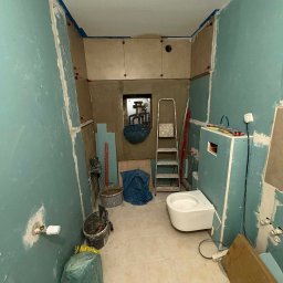 Łazienka w trakcie remontu: ściany z płyt karton-gips, częściowo położone płytki, zamontowana toaleta podwieszana, drabina i narzędzia. Widoczne ślady szpachlowania.