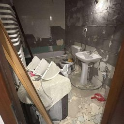 Łazienka w trakcie demontażu płytek, widoczne gruzowisko, umywalka na postumencie, wanna i narzędzia. Przygotowanie do remontu, ciemne płytki częściowo zerwane.